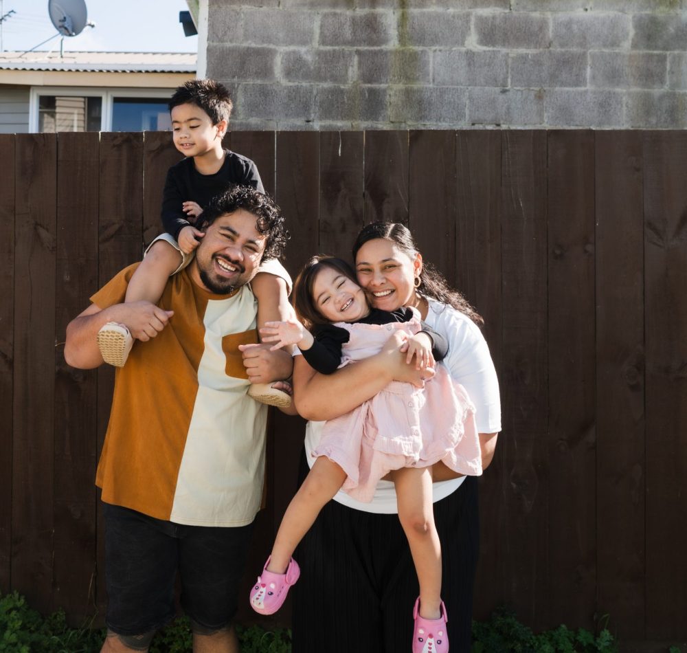 Pacific Islander family of four smiling and having fun standing against brown background.